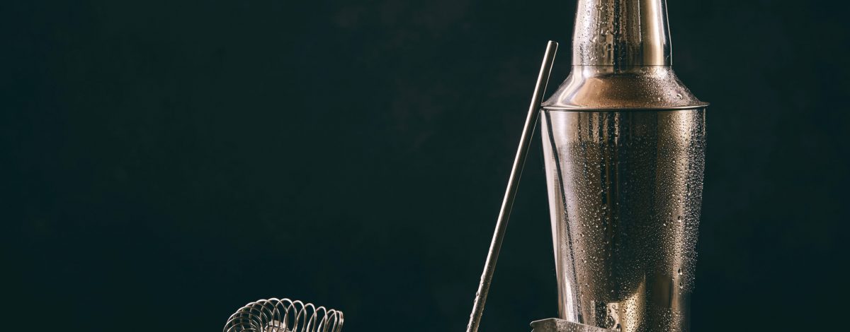 Cocktail shaker, swizzle, tongs and spoon with crushed ice for preparing a summer cocktail beverage on an old vintage wooden table with copy space